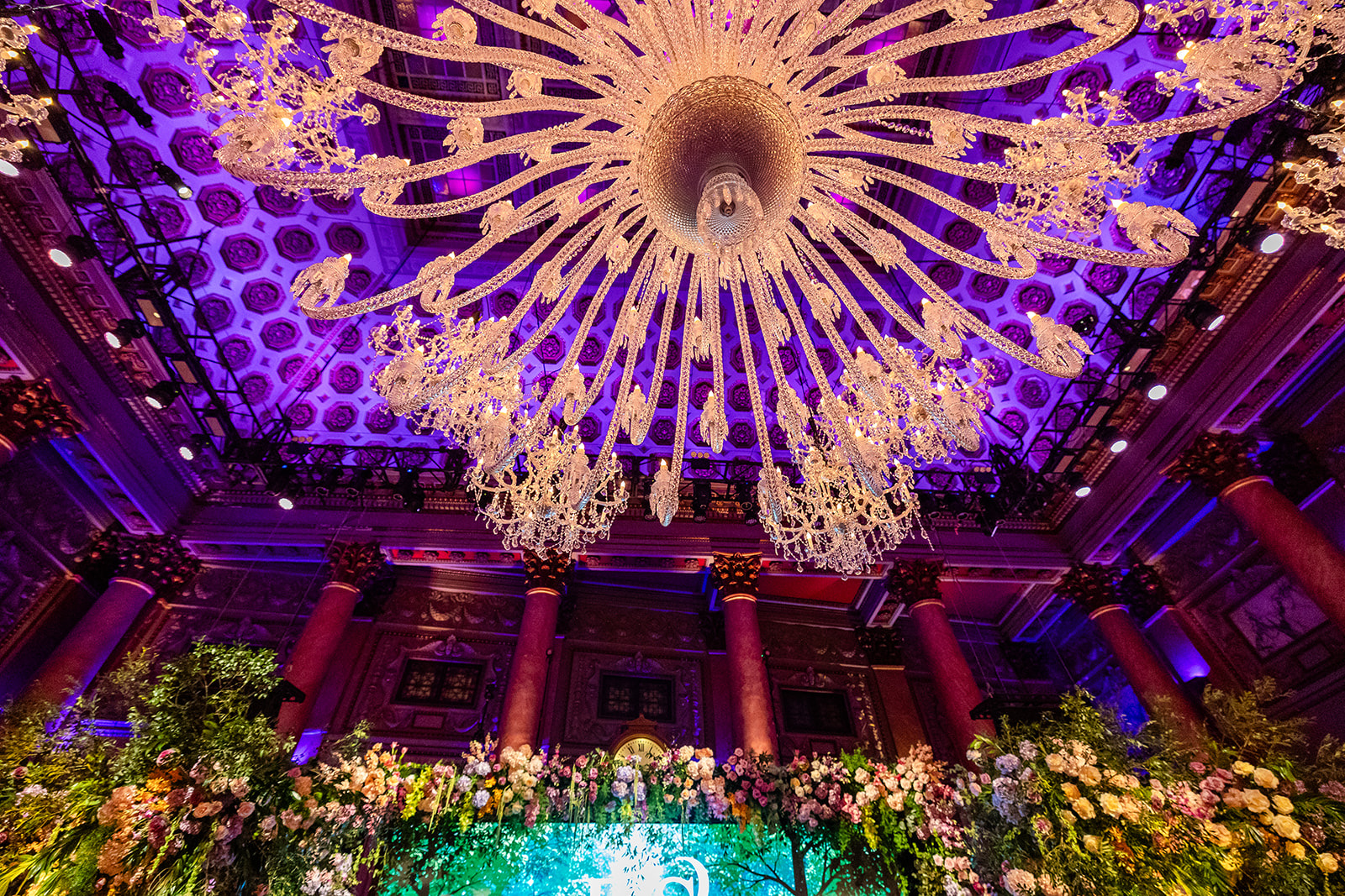 Looking up at the coffered ceiling and central chandelier at Capitale, columns lit in purple below