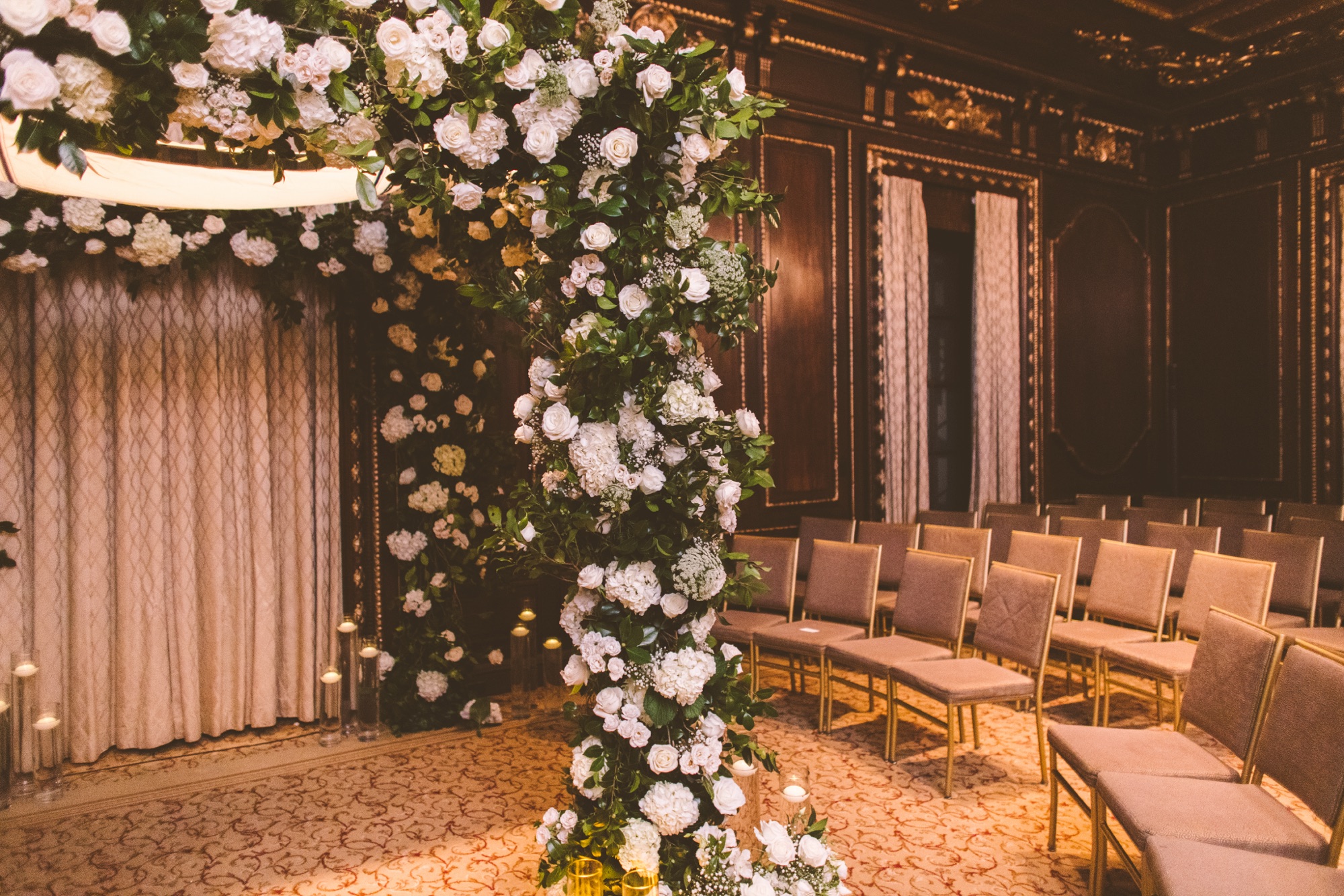 An empty chuppah of white roses and greenery with a tallit canopy above rows of chairs