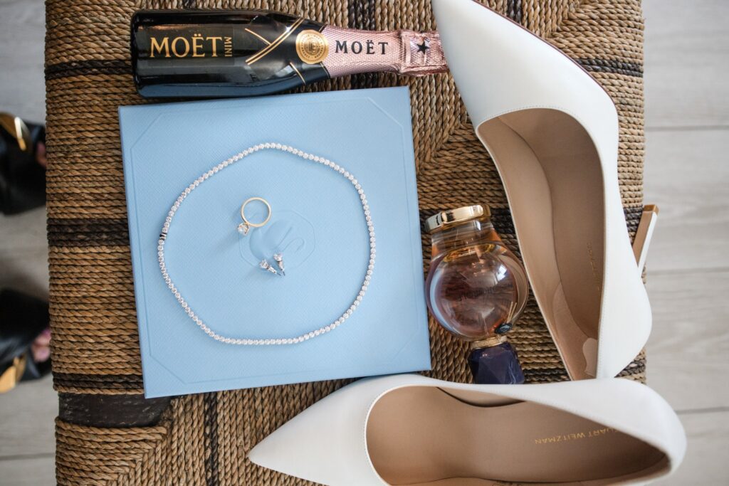 Bridal details laid out before a Deal New Jersey wedding: lace heels, a strand of pearls, a blue jewelry box and a bottle of champagne
